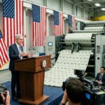 Jerome Powell ยืนพูดตรงโพเดียม พร้อมกับมีธงชาติสหรัฐแล้วเครื่องพิมพ์เงินอยู่ข้างหลัง